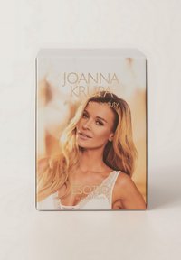 Białe opakowanie z zamknięciem zbliżeniowym przedstawiającym kobietę z długimi blond włosami, ozdobioną koronkowymi akcentami oraz złotym napisem "Joanna Krupa."