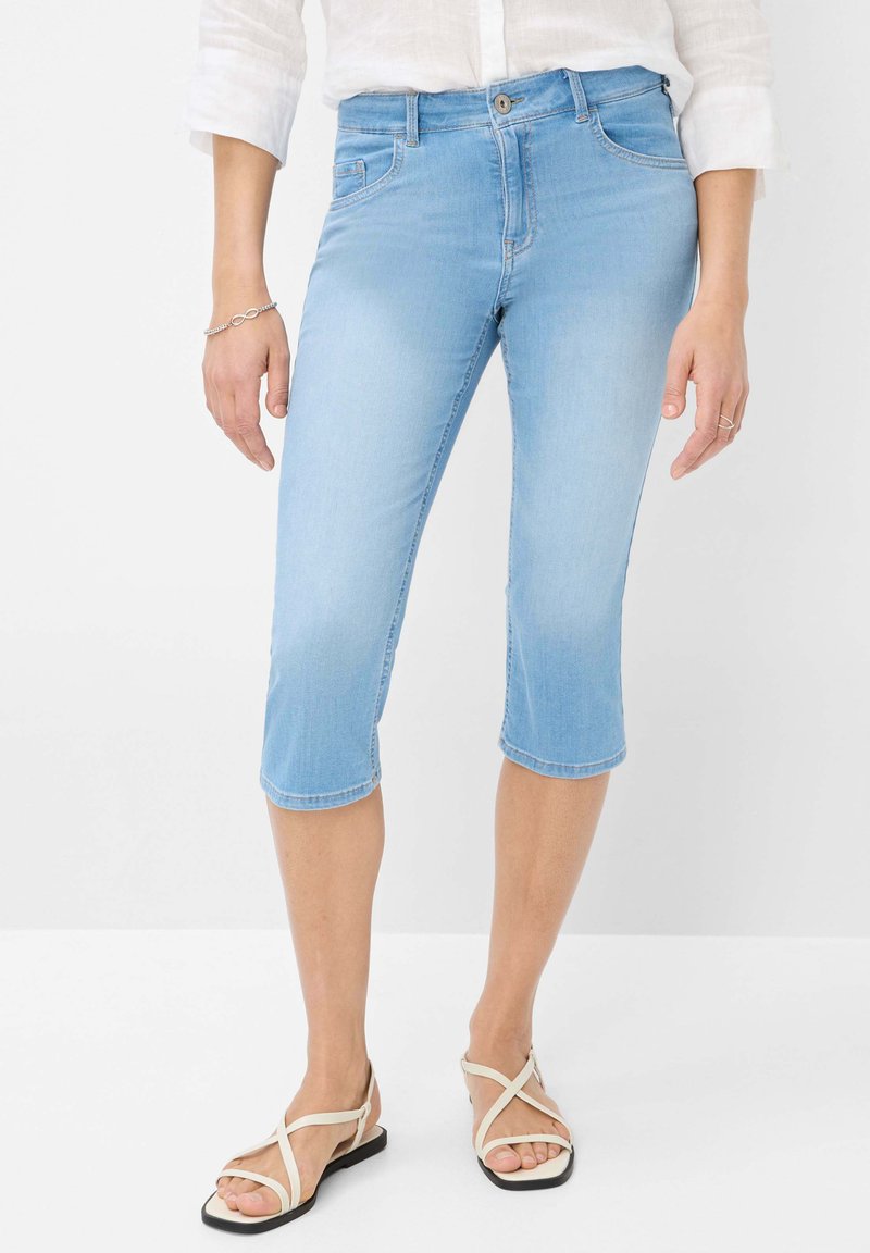 Pantalons capri en denim bleu clair, avec une coupe ajustée, taille mi-haute et ourlets retroussés, associés à des sandales croisées beiges.