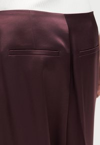 Πίσω μέρος σκούρου μπουρντό παντελονιού με δύο οριζόντιες σχισμές τσέπες, φορεμένο με λευκό ριμπ μπλουζάκι που έχει μπει μέσα.