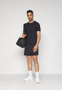 Schwarzes T-Shirt mit kurzen Ärmeln und schwarze Shorts, kombiniert mit weißen Sneakers und Knöchelsocken, während er eine dunkle Sporttasche trägt. Neutrale Hintergrund.