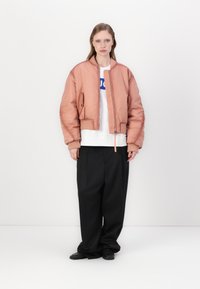 Peach-colored bomber jacket with a ribbed collar and cuffs, zip closure, paired with a white T-shirt and wide black trousers. Smooth texture.