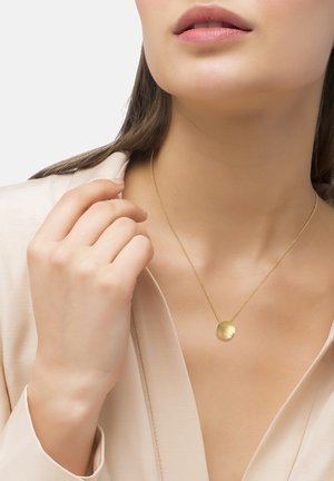 Gouden ketting met een ronde hanger, gladde textuur en kleine accent. Draagt op een slanke ketting, gestyled met een lichtgekleurde blouse.