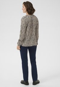 Long-sleeve blouse in a patterned fabric featuring flowing lines in beige and black. Paired with dark blue jeans and black footwear.