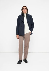 Abrigo peacoat de lana azul marino con botones cruzados, combinado con un suéter de punto blanco. Pantalones marrón claro y mocasines negros completan el conjunto.