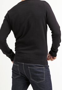 Schwarzer gestrickter Pullover mit Rundhalsausschnitt, gerippten Bündchen und Saum, taillierte Form, getragen mit dunkelblauen Jeans. Rückansicht dargestellt.
