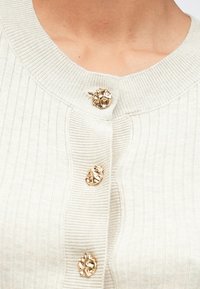 Close-up de um suéter de malha canelada em creme, com um decote largo e três botões de ouro texturizados na frente.