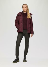 Veste matelassée bordeaux avec fermeture éclair et capuche, associée à un pull sombre, un pantalon noir et des bottes noires imposantes. Texture lisse.