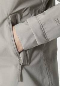 Chaqueta gris claro con una textura suave, que presenta un bolsillo con cremallera y un puño con un acento de logo.