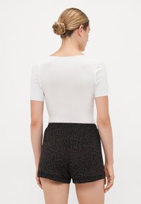 Kvinna med flätad knut som bär en vit kortärmad topp och mörka leopardmönstrade shorts, vänd bort från kameran mot en enkel bakgrund.