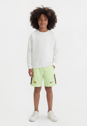 Niño con cabello rizado lleva sudadera blanca, pantalones cortos verde claro con logotipos y zapatillas blancas, de pie contra un fondo blanco liso.
