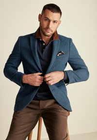 Maßgeschneiderter blauer Fischgrät-Blazer mit braunem Kragen über einem navyfarbenen Polohemd, kombiniert mit braunen Hosen. Ein Taschenquadrat sorgt für das gewisse Etwas.