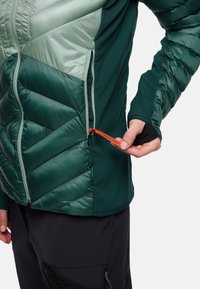 Giacca ibrida verde con un mix di materiali opachi e lucidi, caratterizzata da pannelli trapuntati angolari, una zip laterale e un'etichetta arancione con la scritta "PACK AWAY".