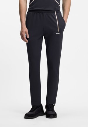 Homme portant un pantalon slim noir avec taille élastique, une petite bande verticale et le logo BOSS sur la cuisse droite, associé à des baskets noires.