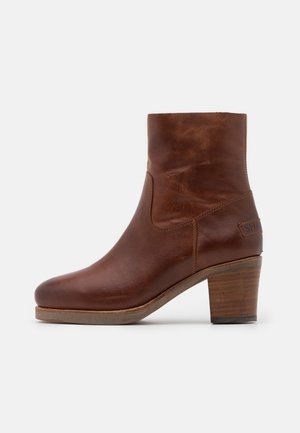 Bottine en cuir marron avec bout arrondi, talon moyen empilé et détail de couture latérale sur fond blanc.