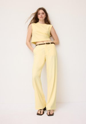 Femme debout sur fond blanc, portant un crop top jaune sans manches, un pantalon large jaune, une ceinture marron et des sandales noires.