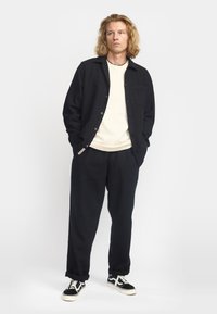 Chaqueta negra de botones sobre una camisa crema, combinada con pantalones negros y zapatillas negras. Presenta un bolsillo frontal en la chaqueta y un corte relajado.