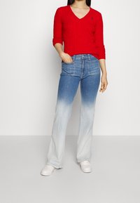 Pull rouge en maille torsadée avec col en V ; jean évasé en denim bleu avec un effet dégradé ; des baskets blanches complètent la tenue.