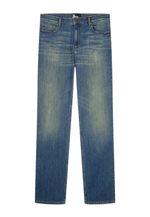 Homme portant un t-shirt bleu marine à manches courtes, un jean bleu délavé, des chaussures habillées noires et des bracelets argentés, debout devant un fond uni.