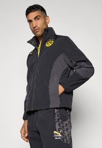 Schwarze Windjacke mit grauen Akzenten, ausgestattet mit einem gelben Logo und Reißverschlusstaschen. Hergestellt aus leichtem Material mit elastischen Bündchen.