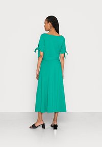 Vestido midi teal com mangas curtas atadas, cintura ajustada e saia rodada. Usado com sandálias de salto pretas. Tecido suave e leve.