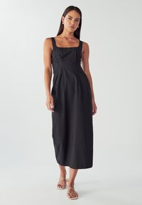 Ženska oblečena v črn midi plašč brez rokavov s kvadratnim izrezom in bež sandali na trakove, ki stoji ob enobarvni svetli ozadju.