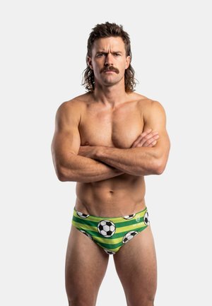 Homme musclé avec une moustache et une coupe mulet se tient les bras croisés, portant un slip de bain vert orné de motifs de ballons de football.