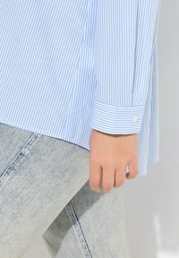 Camisa de botones a rayas azul y blanca con mangas remangadas, con una textura suave, combinada con jeans de mezclilla claros.