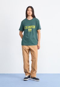 Zielona bawełniana koszulka z żółtym napisem "CHAMPION ATHL. DET.", w zestawie z beżowymi spodniami i granatowymi butami sportowymi na jasnoniebieskiej podłodze.