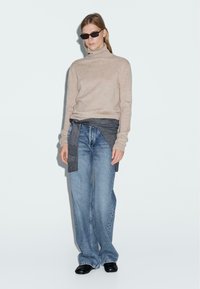 Maglione beige a collo alto, tessuto grigio legato in vita, jeans larghi blu chiaro, scarpe nere, indossato su uno sfondo semplice.
