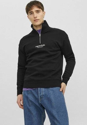 Svart zip-up huvtröja i mjukt material, med hög krage och vit logotyp på bröstet. Kombinerad med ljusblå jeans.