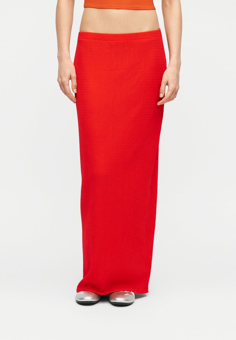 Γυναίκα που φοράει φωτεινή κόκκινη εφαρμοστή φούστα μήκους μέχρι τον αστράγαλο, συνδυασμένη με ασημένια επίπεδα παπούτσια, μπροστά σε απλό φόντο.