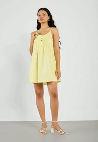 Vestido amarelo sem mangas com um design plissado, apresentando um fechamento frontal com botões e um ajuste solto, combinado com sandálias bejes de tiras.