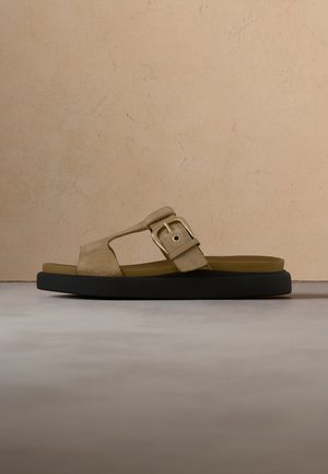 Sandale beige en daim avec une grande boucle dorée et une semelle noire épaisse sur un sol gris et un mur beige texturé en arrière-plan.