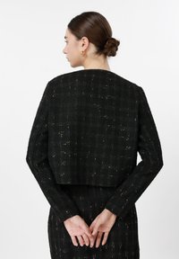 Γυναίκα με σκούρα καστανά μαλλιά σε κότσο, ντυμένη με μαύρο υφασμάτινο σακάκι με διακριτικό καρό σχέδιο, στέκεται με τα χέρια της δεμένα πίσω από την πλάτη.