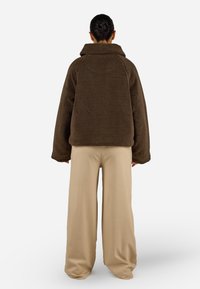 Giacca in pile marrone con colletto alto, vestibilità oversize e spalle scese, abbinata a pantaloni a gamba larga di un beige chiaro.