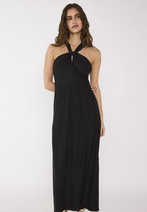 Femme portant une robe noire sans manches à col licou avec tissu plissé, debout avec une main sur la hanche sur un fond blanc uni.