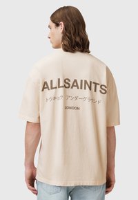 Béžové bavlněné tričko s krátkými rukávy, na zadní straně s velkým tmavohnědým textem "ALLSAINTS" a japonskými znaky, s kulatým výstřihem.