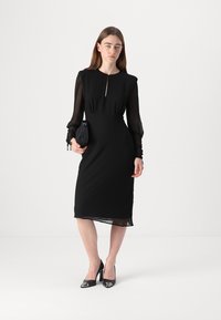 Rochie midi neagră cu mâneci lungi din material transparent, decolteu înalt și detaliu sub formă de cheiță. Material fin cu o siluetă ajustată și tiv de lungime până la genunchi.