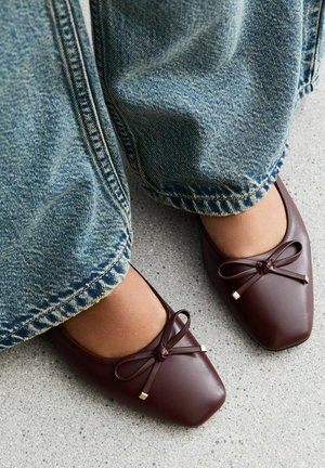 Brown leather ballet flats with front bows worn with wide-leg blue jeans on a gray speckled floor.