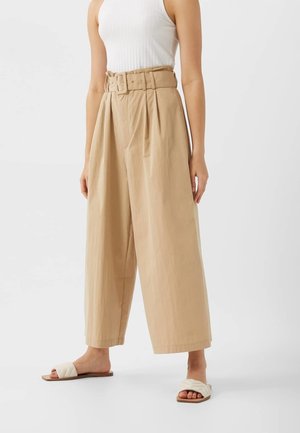 Beige brede broek met een ceintuur in de taille, voorzien van plooien en een gladde stofstructuur. Gecombineerd met witte slidesandalen.