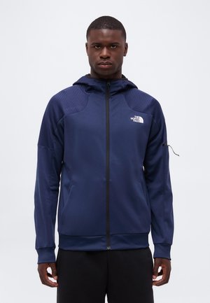 Mann trägt einen marineblauen Zip-Hoodie mit Kapuze und The North Face Logo auf der linken Brust, steht vor einem weißen Hintergrund.