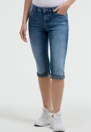 Woman wearing rolled-up blue denim capri pants with leopard print cuffs and white athletic shoes, standing against a plain background.