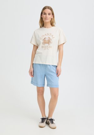 Femme debout portant un t-shirt beige "Coastal Breeze", un short en jean bleu clair et des baskets beiges avec des lacets noirs sur un fond blanc uni.
