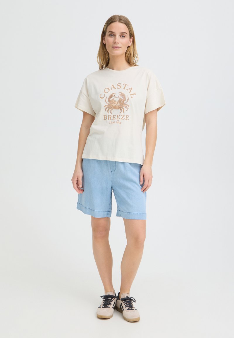 Femme debout portant un t-shirt beige "Coastal Breeze", un short en jean bleu clair et des baskets beiges avec des lacets noirs sur un fond blanc uni.