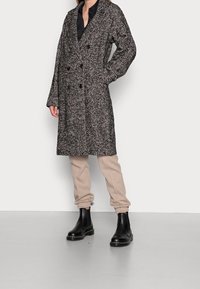 Cappotto doppiopetto in motivo pied de poule nero e bianco, caratterizzato da un tessuto testurizzato, grandi bottoni e tasche laterali.