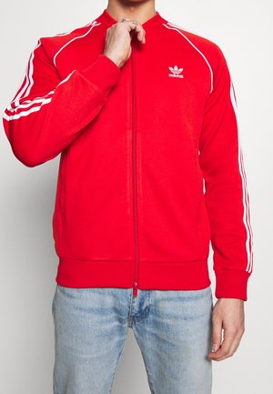 Mann trägt eine rote Adidas-Jacke mit Reißverschluss, weißen Streifen an den Ärmeln und hellblaue Jeans, Hand nahe am Kragen.