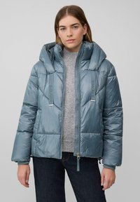 Veste à capuche rembourrée en tissu bleu doux. Équipée d'une fermeture éclair, de logos sur les bretelles et de poignets côtelés. Portée par-dessus un pull gris clair.