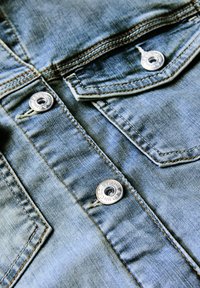 Gros plan sur un tissu en denim bleu clair avec une poche boutonnée dotée de boutons métalliques et de détails de couture contrastés.