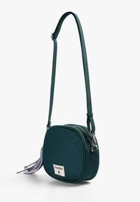 Bolso cruzado verde hecho de tela con forma redonda. Presenta un cierre de cremallera y un borlón decorativo con acentos morados.