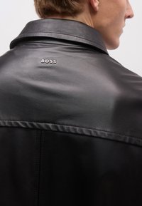 Fekete bőrkabát sima textúrával, gallérral és egy kis, fém "BOSS" logóval a felső háton. Tiszta és modern dizájn.